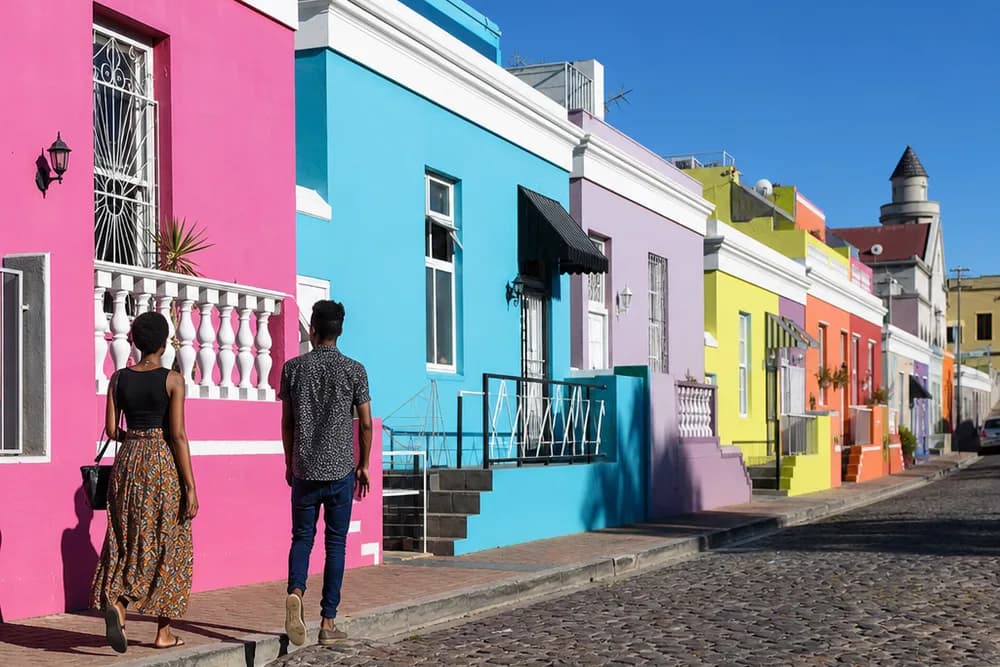 A sunlit cobbled street of brightly painted Bo-Kaap houses with two people walking on the pavement