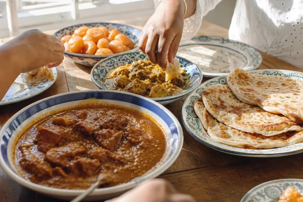 A daylit table of Cape Malay food (curry, roti and koeksisters on ceramic plates) with hands reaching in