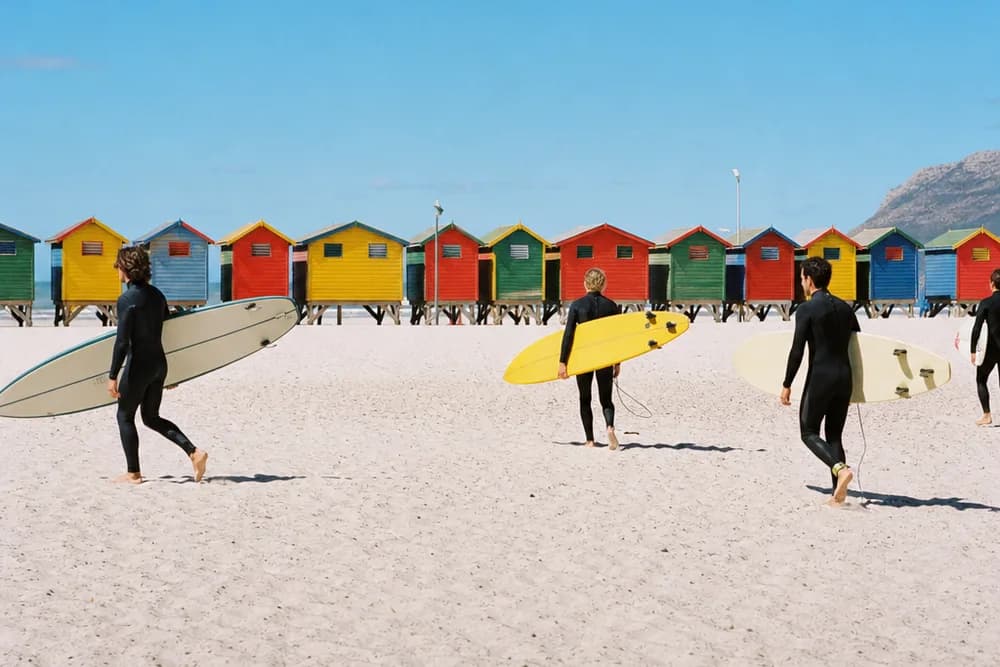 Surfers carrying boards across the sand at Muizenberg, the row of colourful beach huts behind them