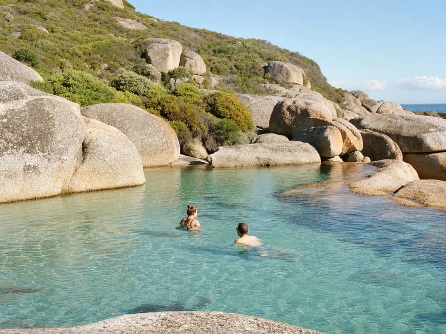 Two swimmers in the clear turquoise water of a rocky tidal pool on the Cape Town coast, granite boulders behind
